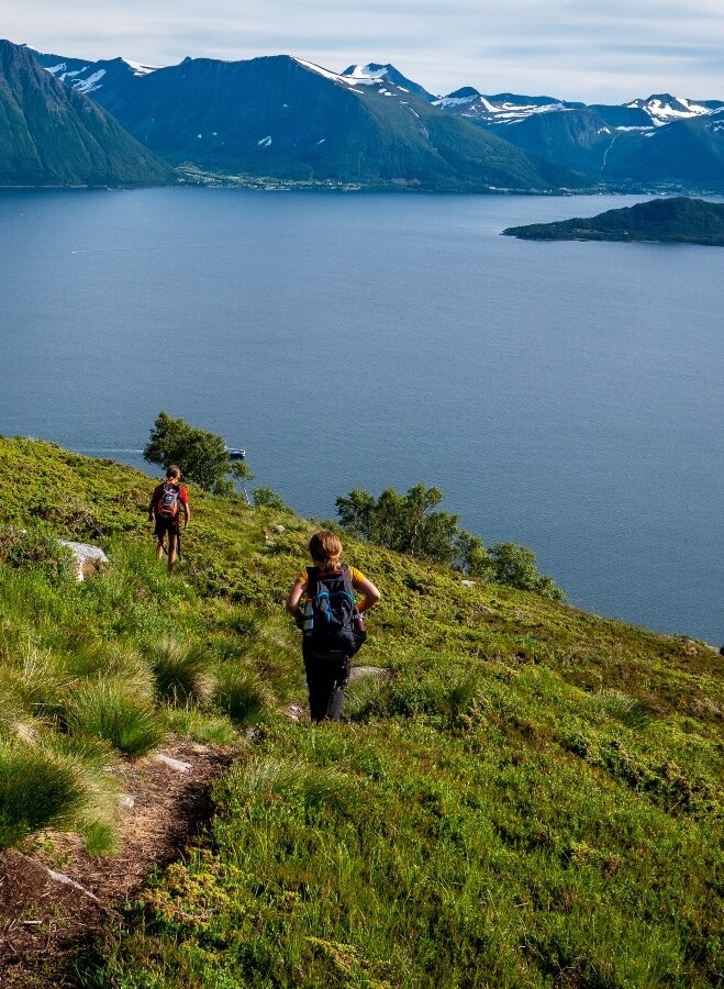 Two hikers trekking a hillside trail above a fjord with mountains in the background.
