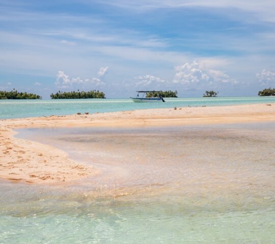 Tropical beach with clear water, a boat, and small islands in the distance under a blue sky.