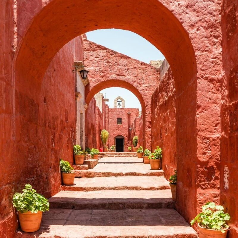 Luxury Peru Tours - Arched pathway in a red-walled monastery with potted plants under bright sunlight.