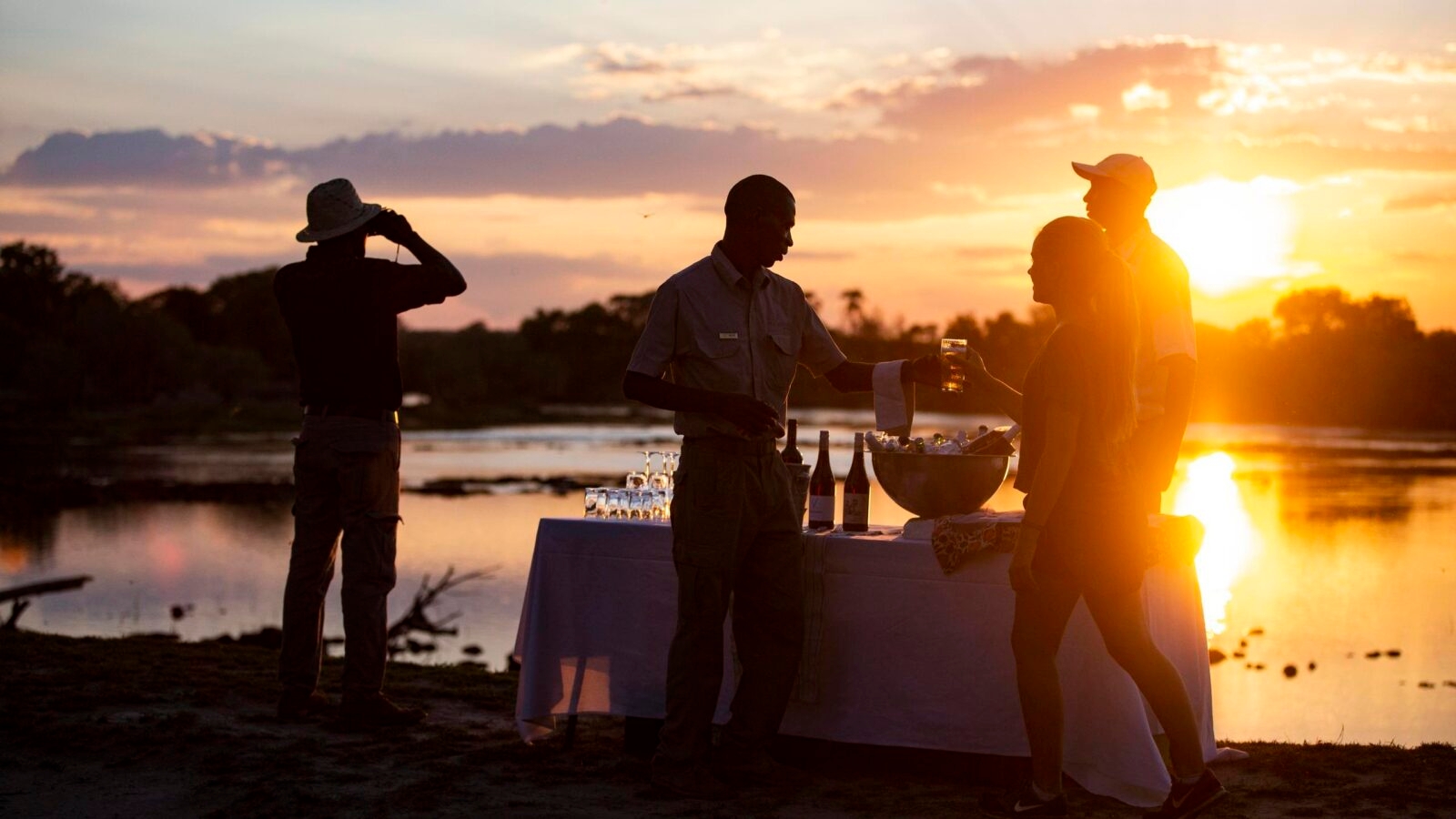Riverbank sunset with a person serving drinks to guests near a table.