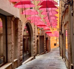 A cobblestone alleyway adorned with a canopy of pink umbrellas, flanked by old stone buildings.