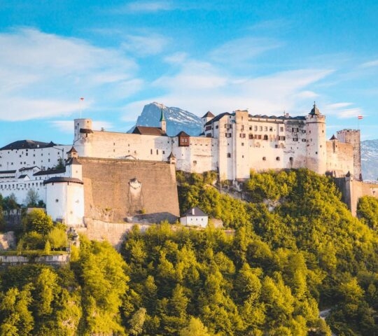 Majestic hilltop castle with white walls, atop a green forested hill with mountains in the background.
