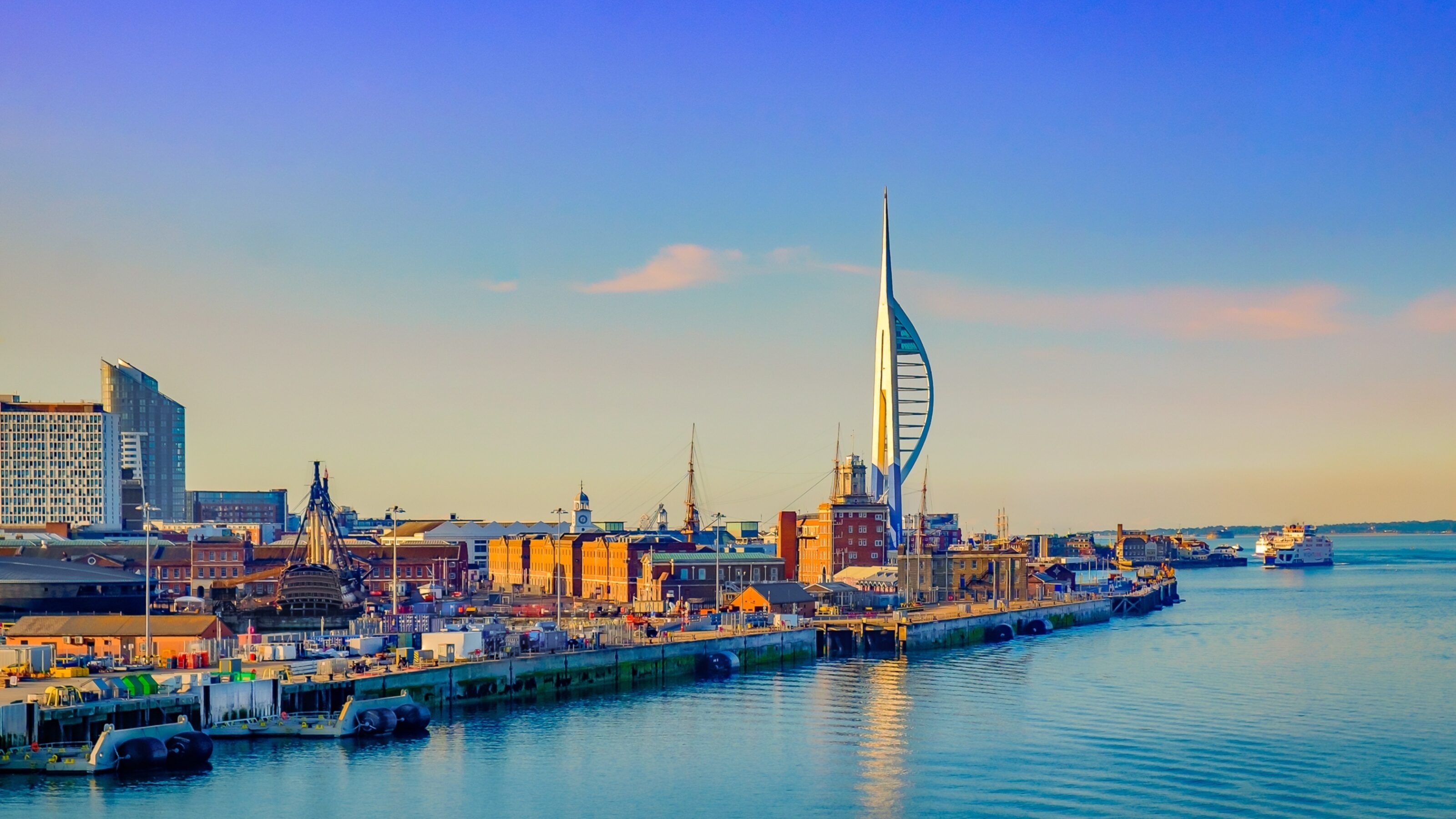 Seaside cityscape during sunset with a tall spire structure and calm blue waters.