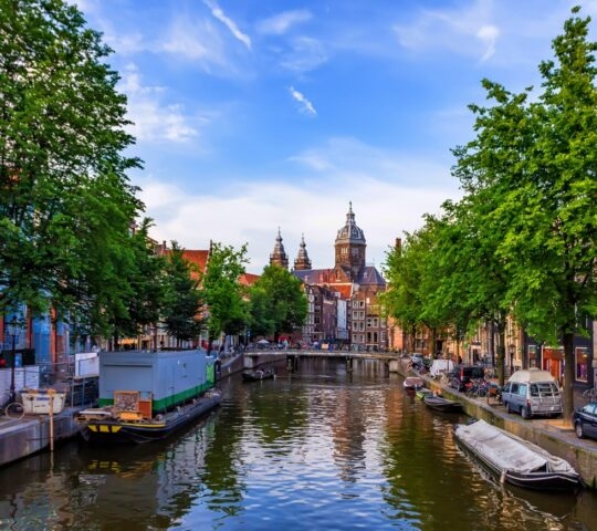 Dutch canal lined with trees and historic buildings under a blue sky with wispy clouds.