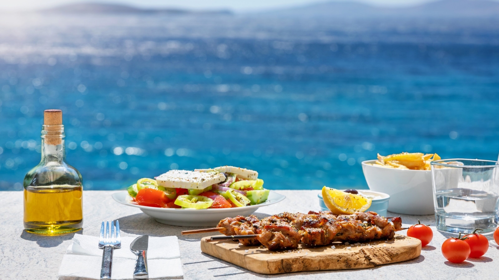 Greek meal with souvlaki, salad, fries, and olive oil, overlooking the sea.