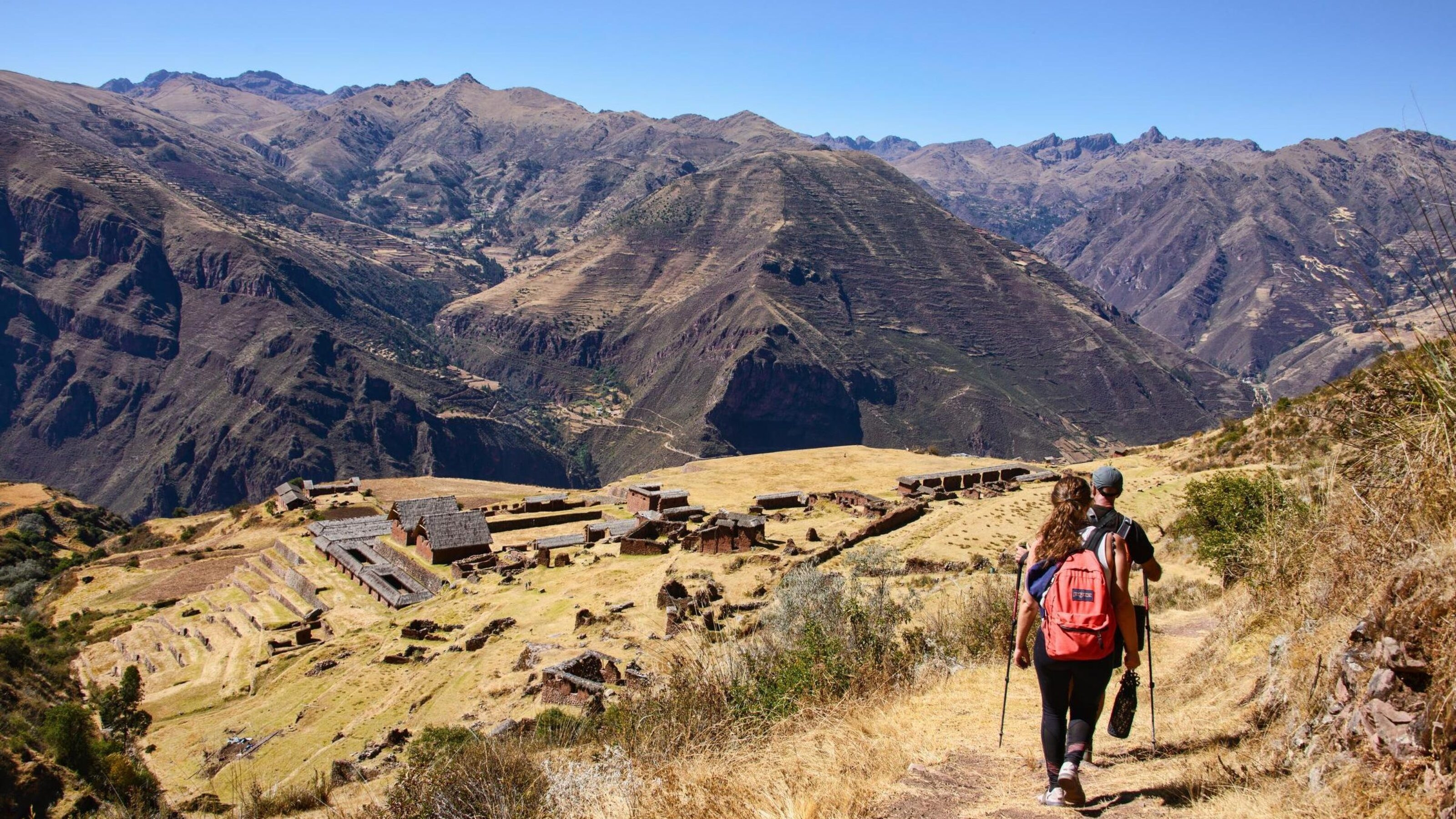 A hiker with a red backpack walking on a trail towards an ancient mountain village with terraces.