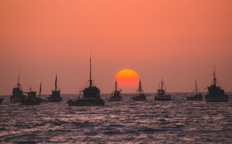 Sunset over the ocean, silhouetting fishing boats against a vivid orange sky.