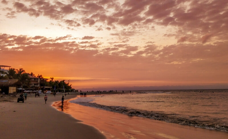 Sunset at the beach with people, reflecting water, and clouds in the sky.