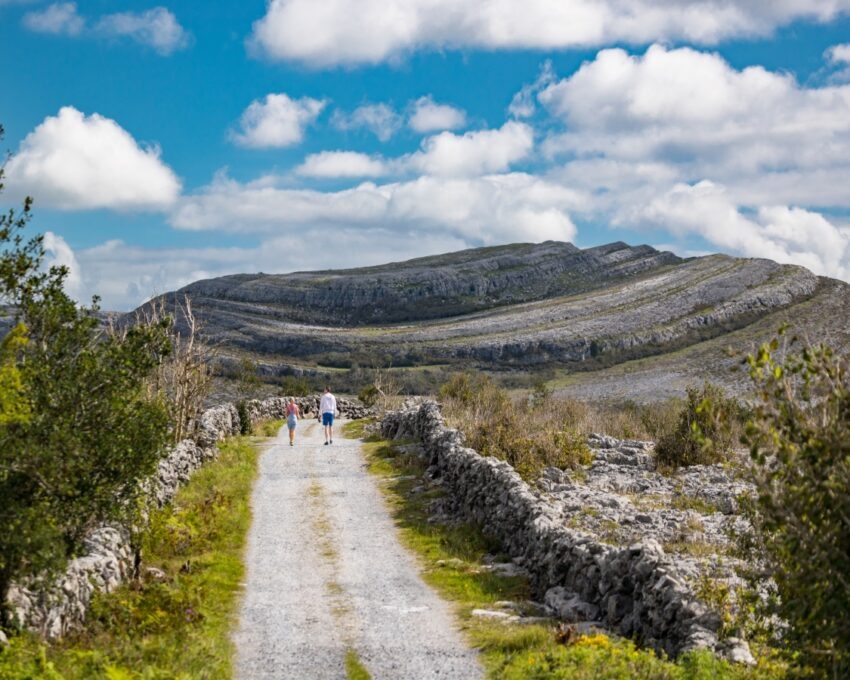 Two people walking on a path with stone walls in a rocky landscape under a cloudy sky.