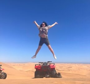 Person jumping above a quad bike on a desert dune under a clear blue sky.