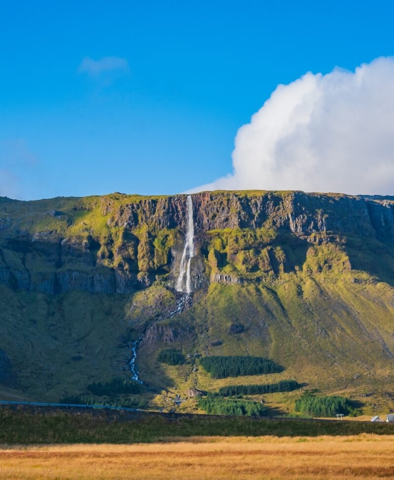 A large rocky mountain under a blue sky, with clouds scattered above. The landscape features a waterfall cascading down the rocks, surrounded by grassy areas and highland terrain.