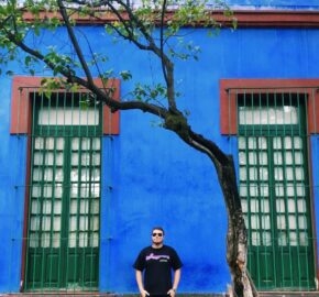 Person in black shirt standing by a bent tree in front of a blue wall with green window frames.