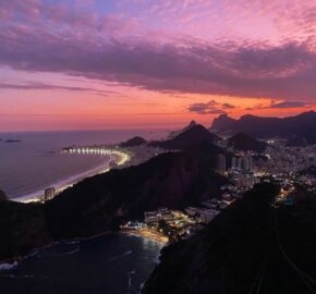 Aerial view of a coastal city at dusk with lit streets and a pink-hued sky.