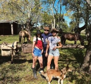 Three individuals in a sunny pasture with horses and a dog, near a saddle on a fence.