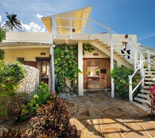A house surrounded by plants with a staircase leading up to the entrance, set against a backdrop of a clear sky and fluffy clouds. The outdoor scene includes a porch and palm trees providing shade, creating a serene atmosphere typical of a luxury hotel