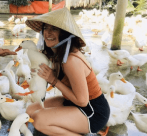 Person crouching among a flock of ducks, wearing a conical hat outdoors.