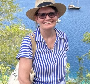 A woman in a blue and white striped dress with a straw hat stands by a lush green area overlooking serene blue waters and distant islands.