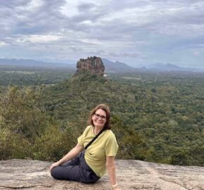 A person sits on a rocky ledge overlooking a lush green landscape with distant hills and a prominent rock formation under a cloudy sky.