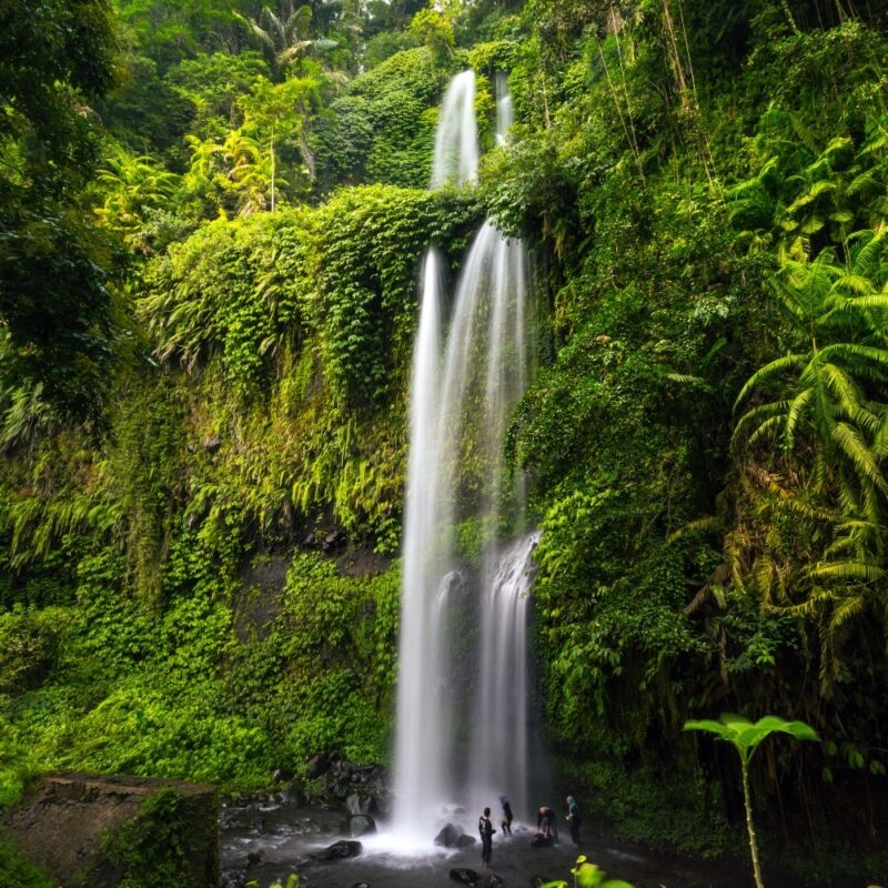 A stunning waterfall cascades down lush, green cliffs, while several people explore the rocky pool below in a tropical paradise.