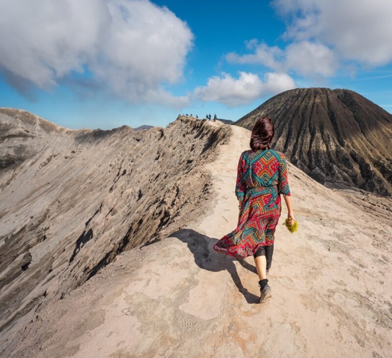 A woman in a colorful dress walks along a rocky volcanic ridge, with a distant volcano and blue sky in the background.