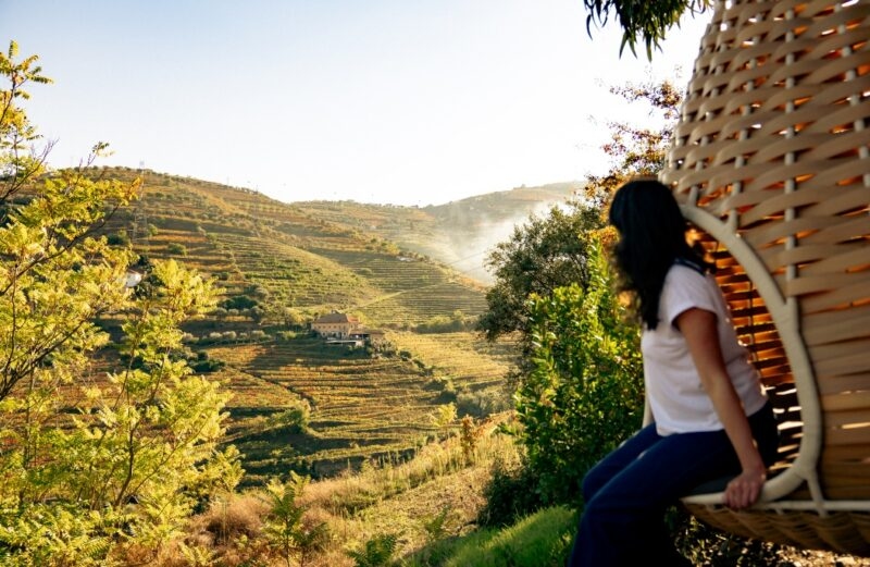 Woman sitting in a woven chair looking at a panoramic view of sunlit terraced vineyards, promoting luxury Portugal vacations.