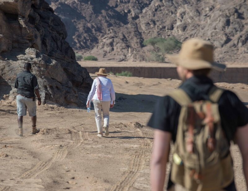 Three people walking across a sandy desert path toward rocky hills during Namibia family tours.