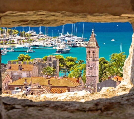 Church and turquoise waterfront view through stone window in Trogir, Croatia.