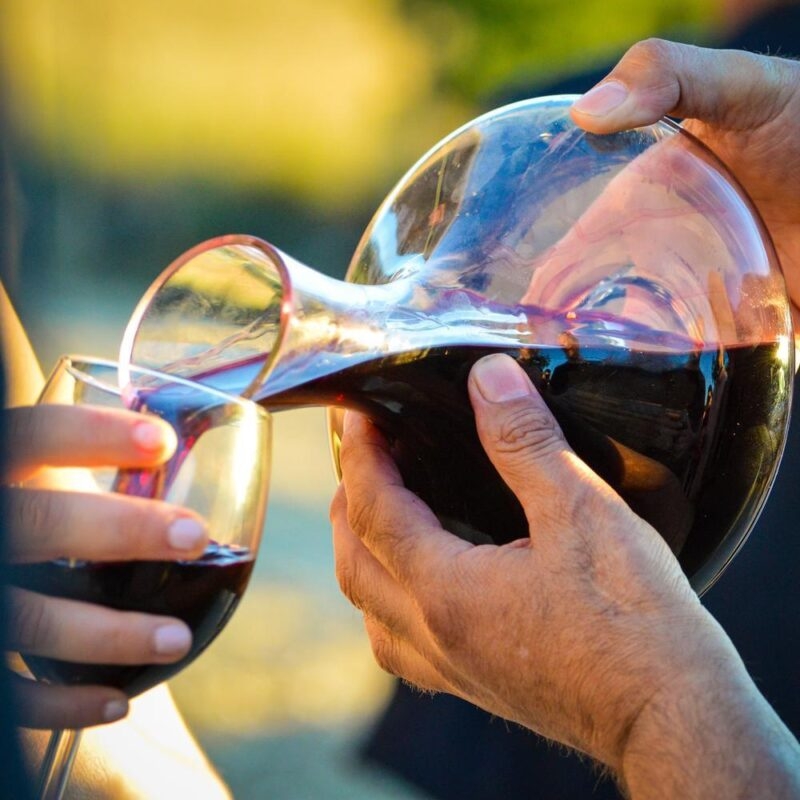 A man's hands pouring red wine from a carafe into a glass for tasting
