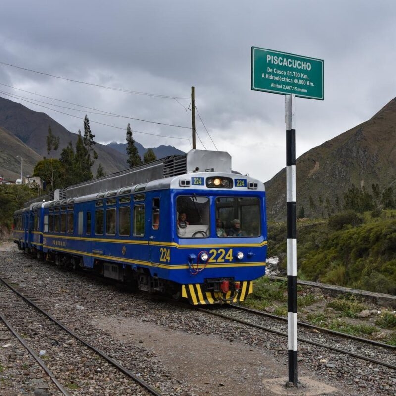 A blue and yellow train on tracks in a mountain valley, passing a sign for "PISCACUCHO," often a part of luxury Machu Picchu tours.