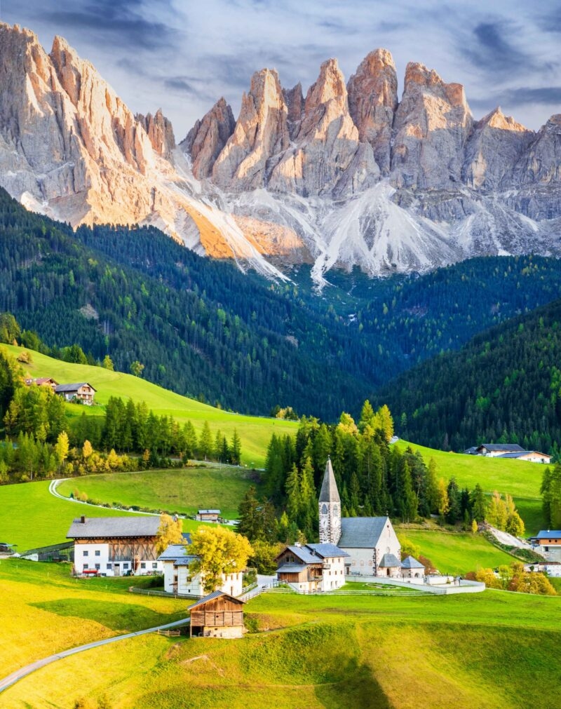 A picturesque village with chalets and a church nestled in a green valley below towering grey peaks.