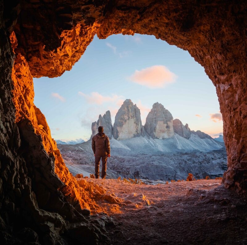 Silhouetted person looking out from a cave opening at a dramatic mountain range during sunset.