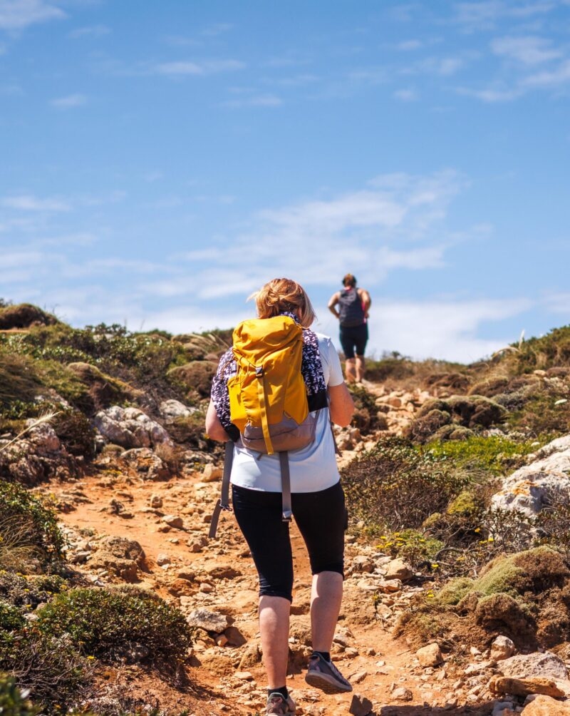 Two people hiking up a rocky, uneven path outdoors under a clear blue sky, a scene from luxury Portugal vacations.