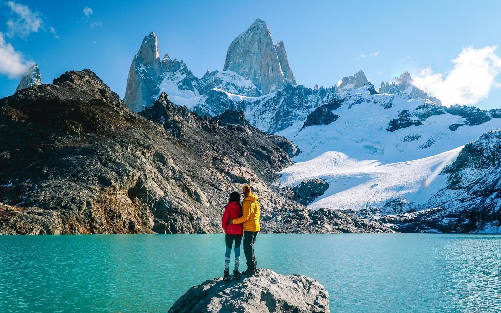 A couple standing at Laguna de los Tres, watching the clear waters and the dramatic granite spires of Mount Fitz Roy in Argentine Patagonia.