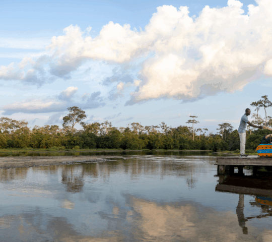 A man serving a woman who is seated on a wooden platform raised above the waters of the Lango baï