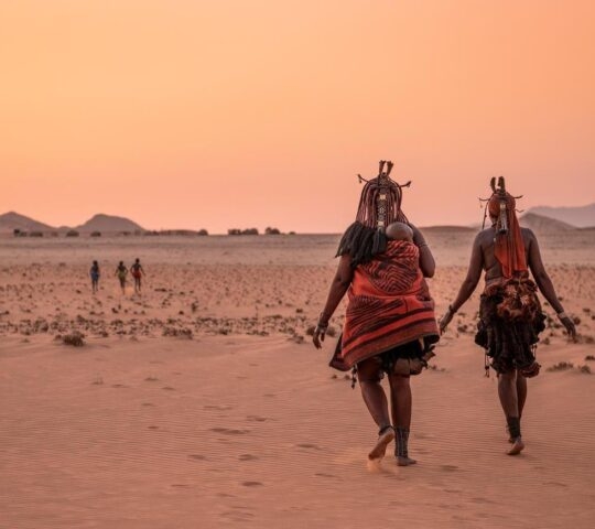 A group of children play in the foreground while a Himba woman with a young child wrapped onto her back walks beside another Himba woman.