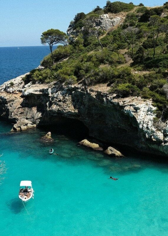 A boat floatin gin turquoise water next to a rocky cliff with trees