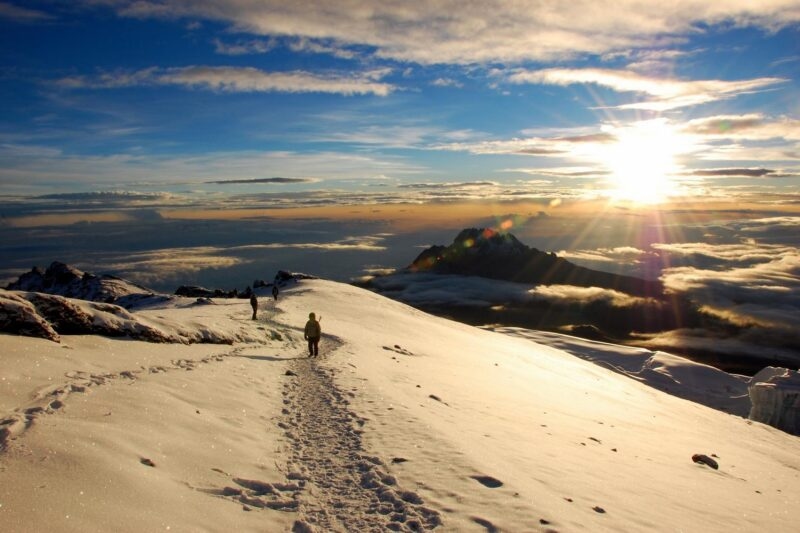 Hikers walk on a snow-covered path on Mount Kilimanjaro at sunrise with clouds below the peak.