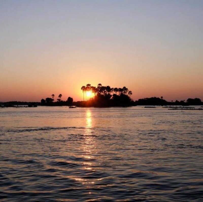 A glowing sun setting behind a small island with palm trees in the middle of a wide river.