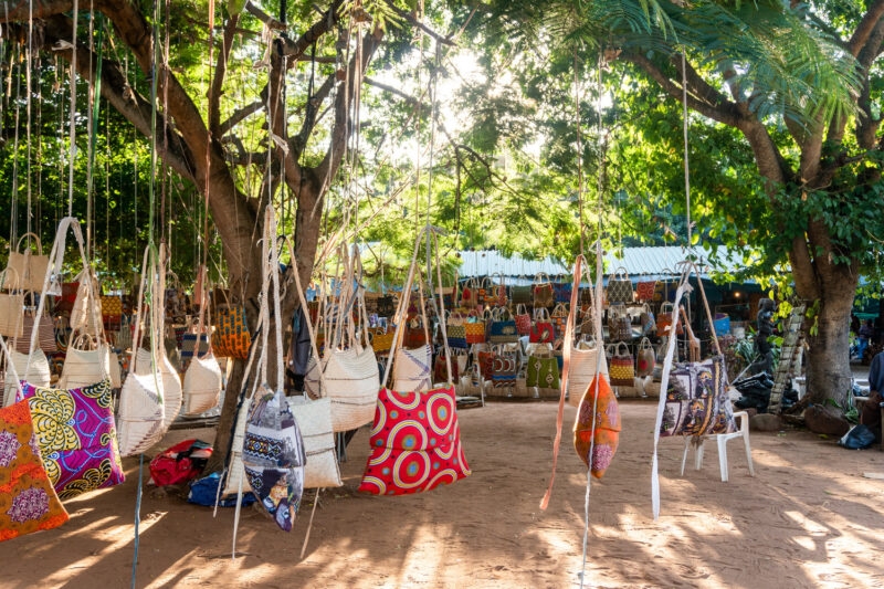 Traditional patterned bags and woven baskets hanging from trees at an outdoor market in Mozambique.