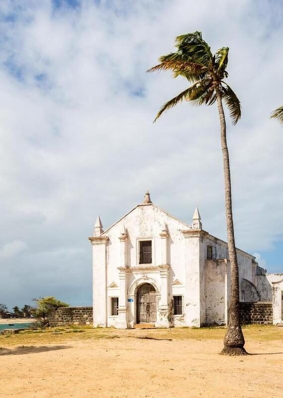 A weathered white stone chapel stands next to a tall palm tree on a sandy beach under a blue and white cloudy sky.