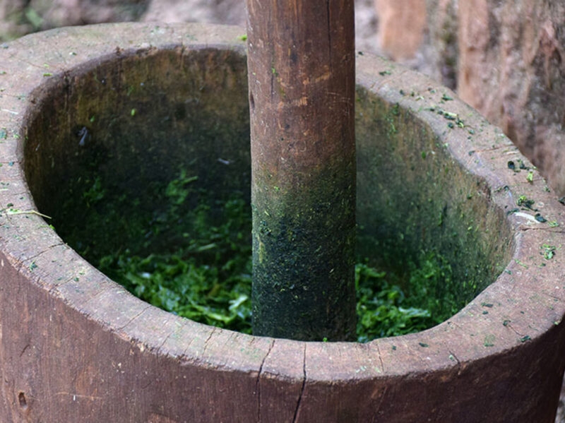 A wooden pestle mashing green leafy vegetables inside a traditional deep wooden mortar in Kigali.