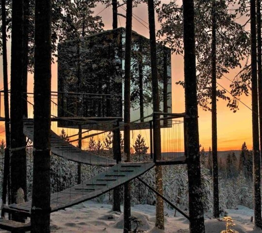 A square mirrored structure suspended in trees reflecting a snowy forest under a sunset sky.