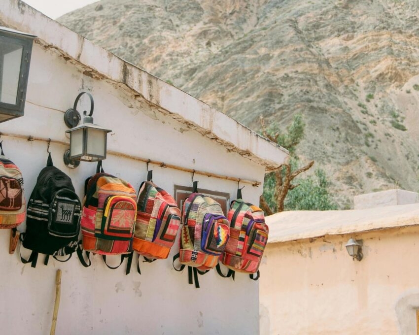 Brightly colored woven backpacks hanging on a white exterior wall under outdoor lamps in a mountain town.