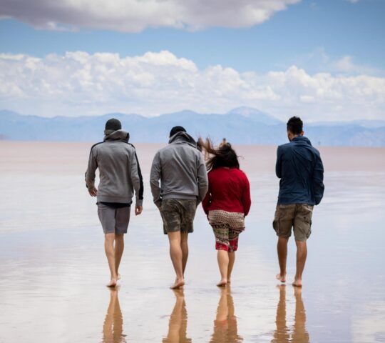Four people walking away on a thin layer of water over white salt flats with mountain reflections.