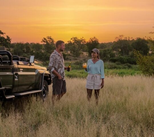 A couple stood next to a safari jeep sharing sundowner drinks at sunset on safari in Africa