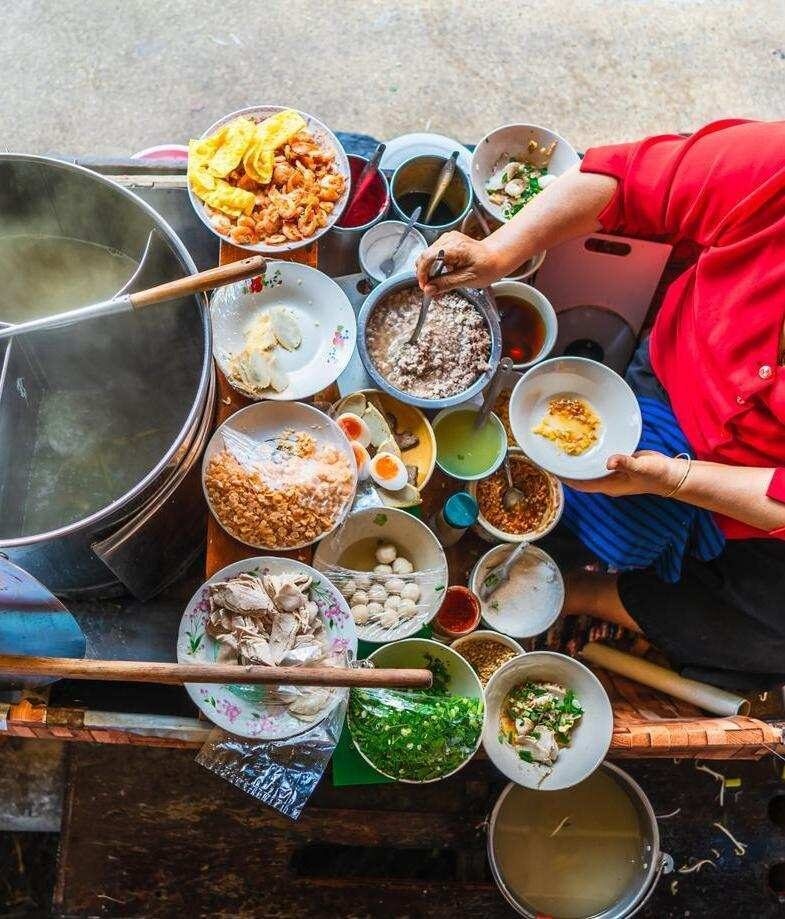 Overhead view of a vendor's workspace on a boat, filled with bowls of noodles, meat, eggs, and herbs.