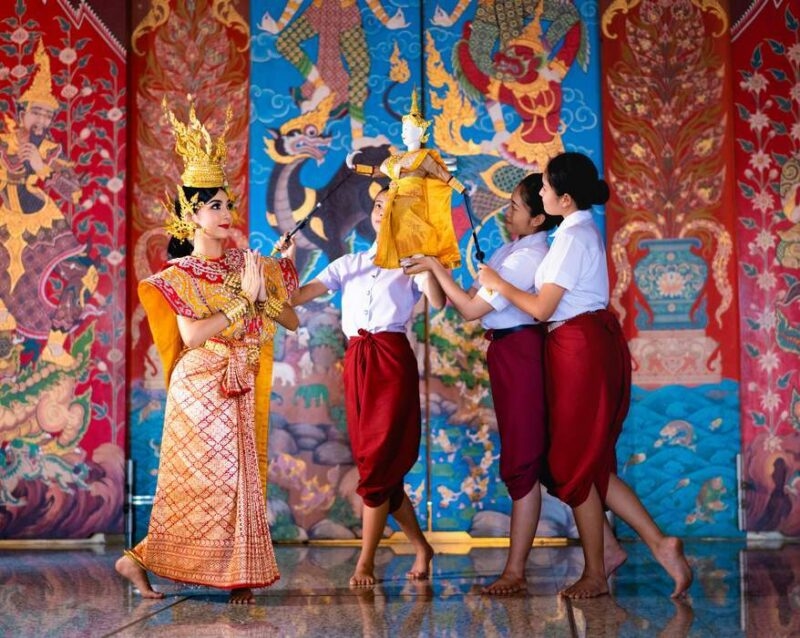 A dancer in gold traditional dress poses next to three people manipulating a Thai puppet against a mural.