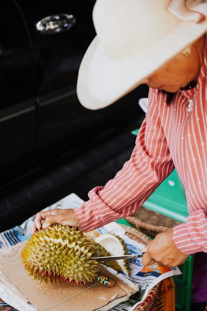 Close-up of a person slicing into a large, green spiky durian fruit at an outdoor market.
