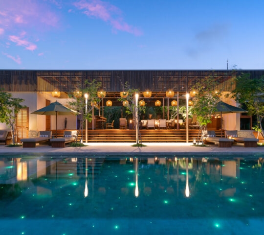 A long swimming pool at dusk with glowing green lights in the water, loungers, and a modern pavilion.