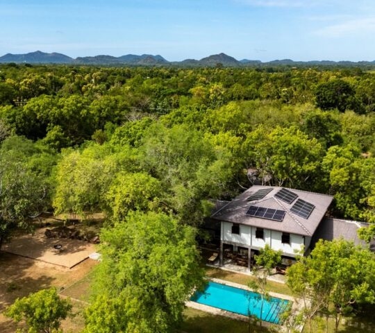 Drone view of a house with solar panels and a blue pool surrounded by a vast green forest.
